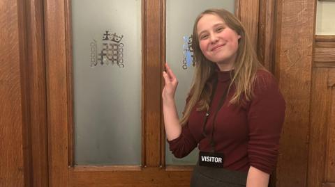 A young woman smiles at the camera. She is standing in front of a door with the Parliament logo on the doors. She is wearing a lanyard with an ID card reading "visitor".