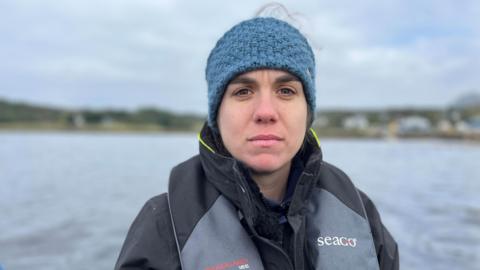 Anna Safryghin wearing a blue woolen head covering, a black jacket and a grey and black lifejacket, with a body of water behind her