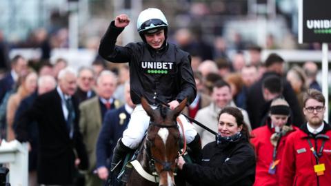James Bowen holds a fist in the air as he is guided while seated on horse Holloway Queen with a crowd behind him at Cheltenham Festival
