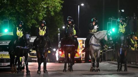 Five police officers wearing riot gear are sitting on horses. They are on a road near traffic lights. There are police vans behind them.