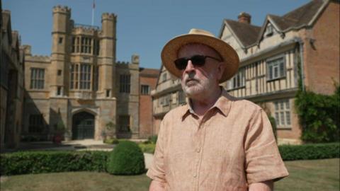 Philip Roberts, a man wearing sunglasses as well as a peach-coloured shirt and a straw hat, is stood in front of an English Tudor building. There is a lawn behind him and a series of low hedges.