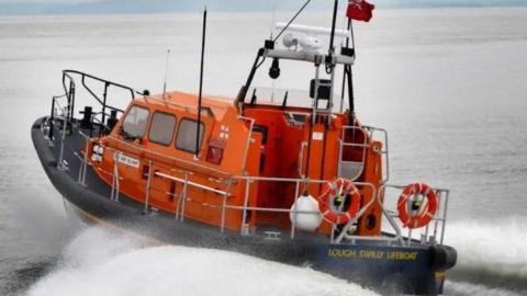 An orange boat with a black trim has the words: 'Lough Swilly Lifeboat' visible.
Waves are visible at the back and side of the boat. 