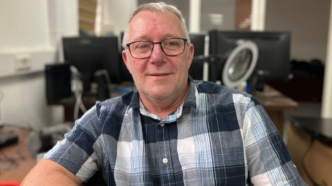 A man in a short-sleeved chequered shirt, with a number of computer screens behind him. The man has short grey hair, and wears dark framed glasses.
