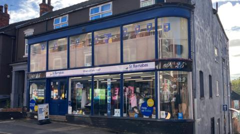 The outside of a blue charity shop called 'St Barnabas'. The shopfront has a navy frame and large glass windows both up and down. Inside there are mannequins clothes and shelves with bric-a-brac in the bottom windows. There is a white sign on the pavement outside. To the right, there is a black sign reading No 25. To the side of the shop are houses, and a large wall painted grey.