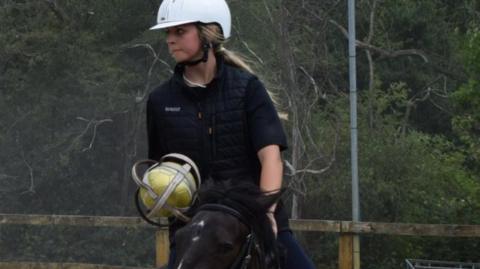 A young woman on a black horse holding a yellow ball wrapped in a metal casing with handles. She is wearing black clothing, a white helmet, and has long blonde hair. She is gazing to the right and in the background are trees and a brown fence.