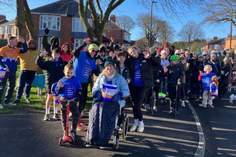 Janine Turnbull in her wheelchair surrounded by her family and dozens of neighbours of all ages. They are filling the street on a sunny day with houses and trees behind.