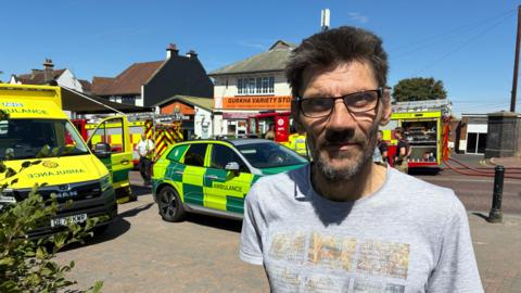 A man with a beard and glasses wearing a grey t-shirt is standing in front of ambulances and fire engines.