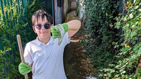 Eight-year-old Jackson wearing sunglasses, white polo top, black shorts and green gardening gloves is standing in an alley way with glass and rubbish. He is holding a brush and giving the thumbs down. There is a metal fence behind him with overgrowing leaves on both sides of the path.