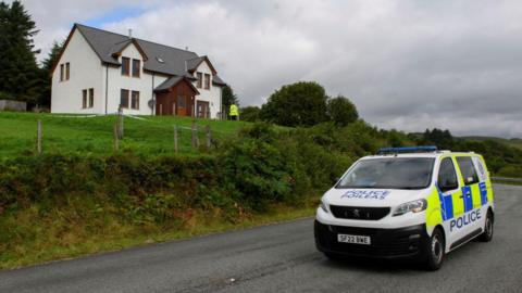 A police van is parked on a road below the two-storey house. There is a police officer near the door.