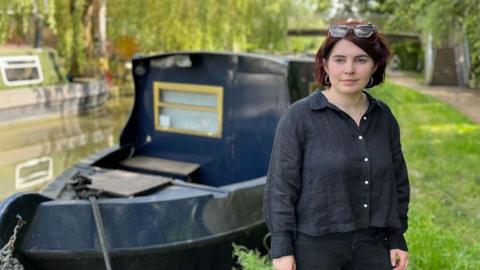 ˿ reporter Clodagh Stenson stands on the bank of the Oxford Canal. She is wearing a black shirt and black trousers, with sunglasses propped on the top of her head. She is standing on a grass bank with a bridge over the canal in the background. Directly behind her is a navy blue narrow boat with a yellow window