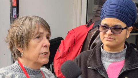 Woman on left wearing a grey jumper, has brown hair and a red lanyard talking into a microphone. Woman on right wearing a hijab, glasses, grey jumper and black cardigan listens in.