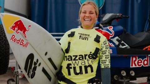 Robyn wearing a bright yellow top over a black wetsuit, smiling at the camera. She holds a Red Bull surfboard in front of a jetski which sits on a trailer. 