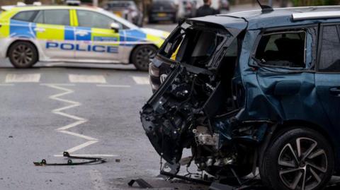 The back of a car following a crash. In the distance a police car is parked behind police tape