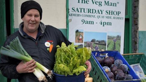 Bry Radcliffe holds leeks, and a tub of leafy greens. He wears a dark jacket and hat and stands next to green boxes full of purple cabbages and pale parsnips. A sign behind him reads 'The Veg Man' and explains the opening times of his Saturday shop.