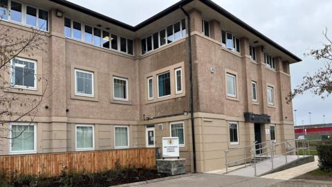 The entrance to Cumbria coroner's court building. The entrance is at the left-hand side of a three-storey building, with beige cladding on the ground floor and shelldash on the upper floors. There is a sign for the coroner's court in front on a ramp leading to the door.