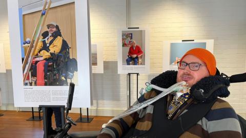 A man, who is paralysed from the neck down and in a wheelchair with breathing tubes attached to him, smiles as he sits beside a photograph of himself with text underneath at an exhibition. He is wearing a striped jumper and a neon orange beanie.