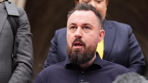 Martin Hibbert sits speaking. A media microphone is in front of him and people stand behind him. 