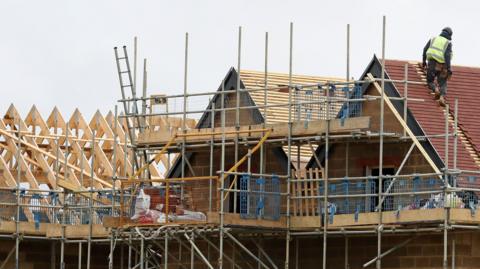 A general view of new homes being built. The roofs are covered in scaffolding. A lone worker wearing a hard helmet and high vis jacket works on one of the roofs.