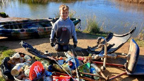 Logan Currie, 7, kneeling in front of a pile of rubbish he and his father collected from a pond. There are bags of cans and a car bumper. He is kneeling next to a camouflaged kayak which his dad used to collect floating rubbish.