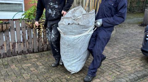 A man in a navy boiler suit, wearing black boots, hold a large white sack with both his hands, which are protected by grey gloves. He stands to the right of the bag. To the left of the bag is a man in a black hoodie, black trousers and boots. They walk in front of a dark-grey fence, on a grey brick driveway. 