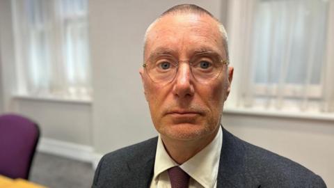 Anthony May in a suit, wearing glasses looks straight at the camera.