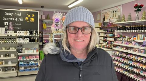 A blonde woman wearing glasses and dressed in a grey coat and a woolly hat in front of a store, with soap and incense on the shelves behind her