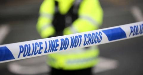 Police officer stands behind police tape reading police in blue on white background