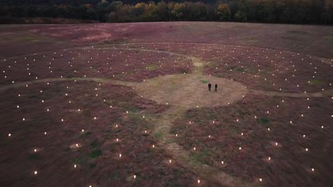 A drone image of two people dressed in dark clothing, who are seen from very far away in a clearing. Around them is a large swirling artwork, lit by hundreds of lanterns spreading out in every direction. There are curved paths running through the installation. 