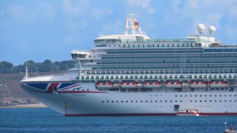 This is a photo of a large cruise ship in the sea in front of some land. The cruise ship is white with the Union Jack flag on the front of the vessel. It bears the name 'Ventura'.