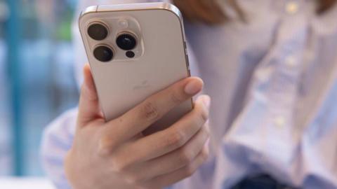 A young woman wearing a light blue, stripy shirt shown in a blurred backdrop holding an iPhone 16 Pro.