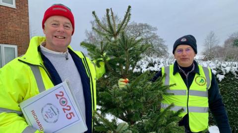 Peter Chapman and Richard Raymond stand next to a Christmas tree. They are smiling and wearing hi-vis clothing and woolly hats. There is snow on the ground and on some bushes behind them.