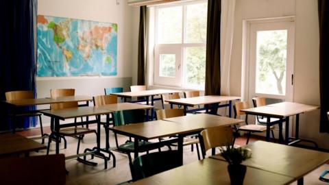 Photo of empty classroom with map of the world on the wall at the back. The classroom is tidy and all the chairs are tucked into the tables. There are large windows with trees showing on the far side. 
