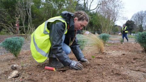 A female volunteer with french braids, wearing a hi vis waistcoast, rainproof jacket and gloves, plants a bulb in the new garden
