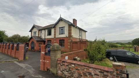 A brick wall in front of a large detached red brick house which used to be a pub.