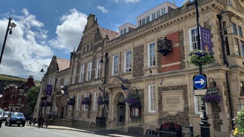 A view of the outside of the Douglas Council buildings on a sunny day. It has an ornate frontage and is decorated with hanging baskets.