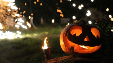 A carved pumpkin sitting on a flat stone outdoors at night. The pumpkin has a wide, triangular-eyed face with a large, curved smile, and it is illuminated from within by a warm orange glow.
Next to the pumpkin, on the left side, there is a small metal torch or lantern with a visible flame. In the background, numerous bright sparks and glowing dots are scattered across the dark scene, likely from fireworks or sparklers.