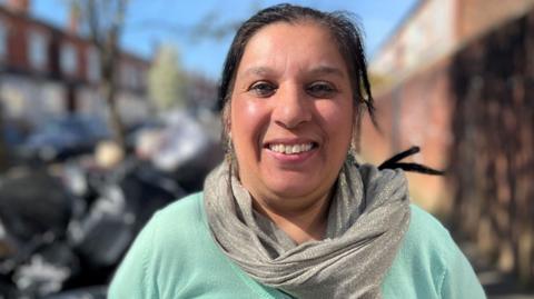 A woman stands on a street on a sunny day. She is wearing a pale green top and a silver scarf. She has straight black hair that is tied back.