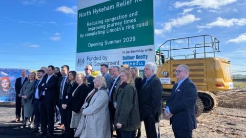 A group of about 20 people, all formally dressed, stand on a building site in front of a large sign reading: "North Hykeham Relief Road, reducing congestion and improving journey times by completing Lincoln's ring road, opening summer 2029, funded by UK government, in partnership with Balfour Beatty." A large yellow, tracked construction vehicle is parked behind the sign and is partially visible.