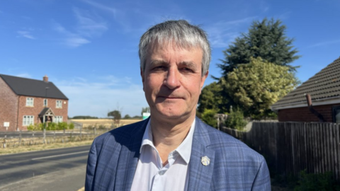 Grey haired man wearing a blue checked jacket and a pale blue shirt with a collar, he is also wearing a pin badge on his lapel. He is standing in front of a road to his right and a brown fence to his left. There is a house behind him across the road and a field. It is a sunny day with blue skies