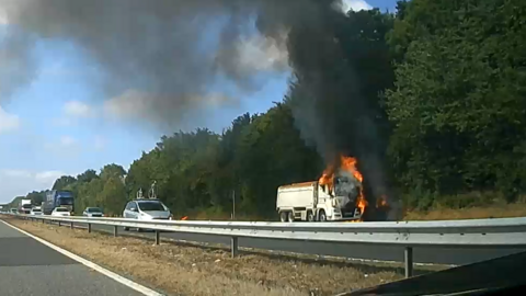 Dashcam footage of a white lorry on fire beside the road. The cab is engulfed in orange flames, with a large plume of black smoke going up into the sky and drifting over the road. There are a number of cars and lorries passing. 
