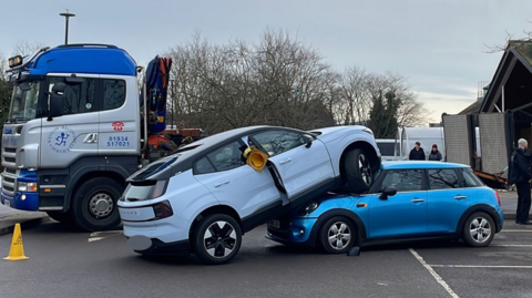 A white car mounted on a blue Mini in a car park, with a recovery truck behind it.