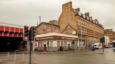A Glasgow street, with a pub on the corner, and a large sandstone building to one side. Just behind it is a railway bridge.
