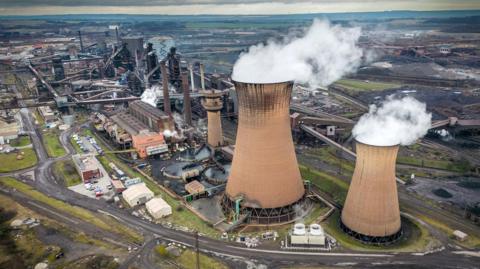 Steam comes out of huge towers in a wide shot of Scunthorpe steel works against a cloudy sky in 2023.