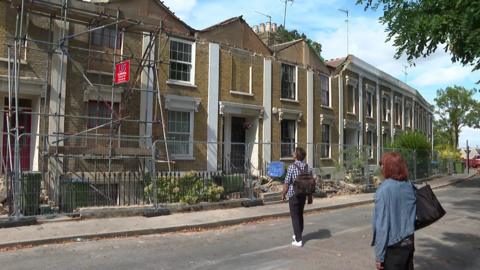 A row of terraced houses on Woodland Terrace in Charlton behind temporary fencing after part of the roofline and frontage collapsed. Two people stand outside the properties, where scaffolding has been erected and debris is visible along the pavement.