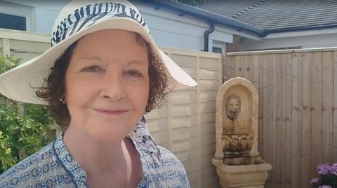 Lesley Middleton has a broad brimmed sunhat, flowered top and red hair. IN the bsackground is a bush and fountain.