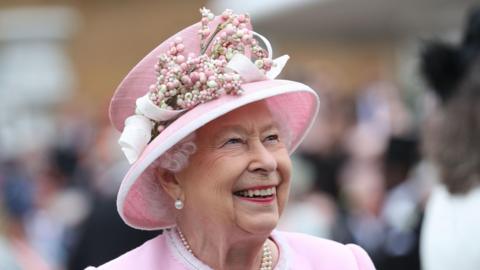 An image of Elizabeth II smiling. She wears a pink jacket and pink hat with a pearl necklace and earrings.