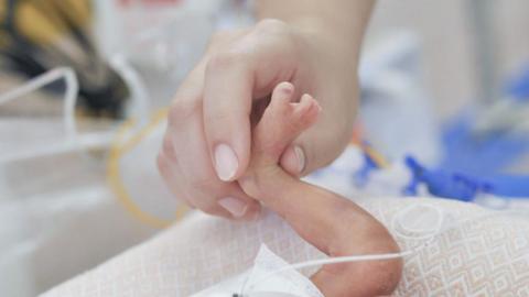 A hand holding a premature baby's foot in a medical setting, with wires visible