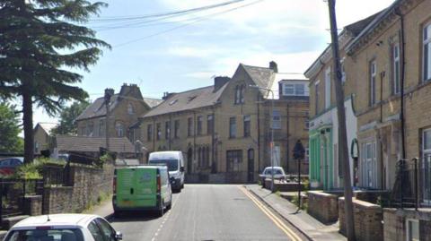 A street in central Bradford, with two and three-storey residential buildings. Cars and vans are parked on the left side of the road, with double yellow lines on the other side. 