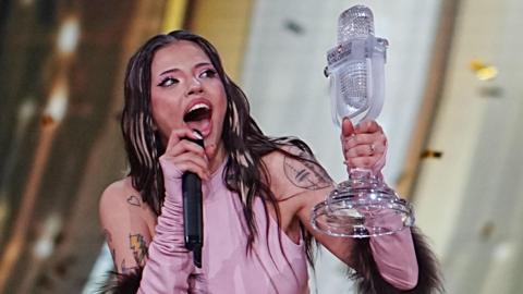 Dara is holding Eurovision's glass microphone trophy after winning the competition in Vienna in May. She is wearing a pink top with her shoulders uncovered, showing several tattoos. She has blonde highlights in her otherwise dark hair and is open-mouthed with excitement. She has pink eyeshadow with dark eyeliner and thick eyelashes. She is talking into a black microphone.