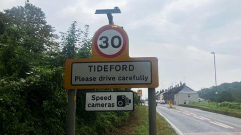 A road speed limit sign which says "30", and, below it 'Tideford, please drive carefully". Below that is a sign saying "speed cameras" and a black and white graphic of a camera. Beside the sign on the right is the road.